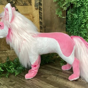 May include: A pink and white plush toy in the shape of a wolf with long white fur and a pink tail. The toy is standing on a wooden surface.