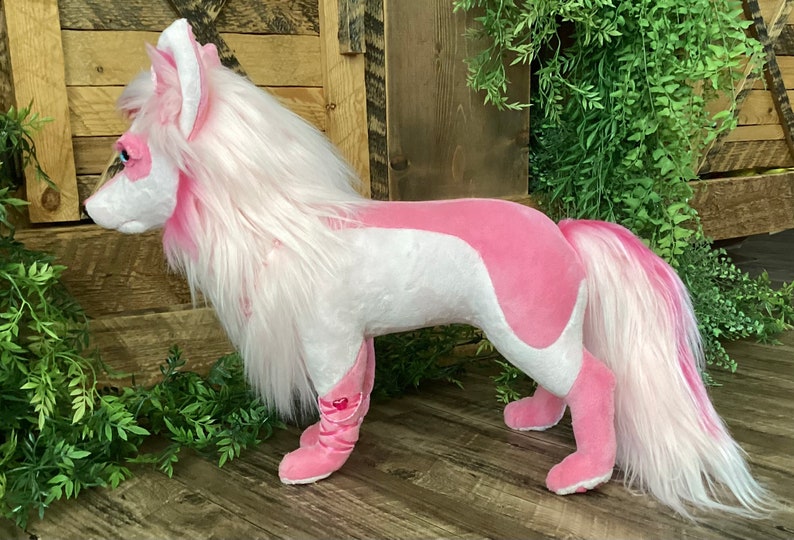 May include: A pink and white plush toy in the shape of a wolf with long white fur and a pink tail. The toy is standing on a wooden surface.