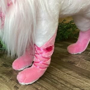 May include: A white plush unicorn with pink boots and a pink beaded necklace. The unicorn is standing on a wooden floor.
