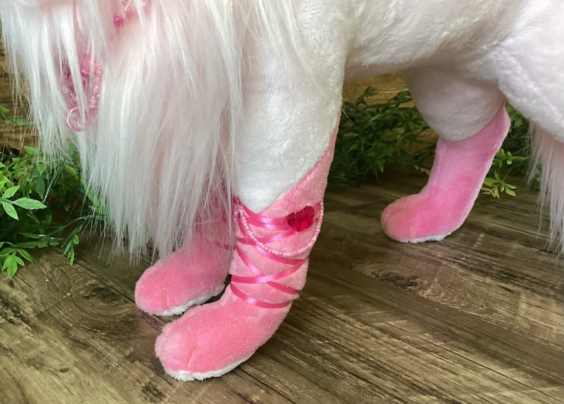 May include: A white plush unicorn with pink boots and a pink beaded necklace. The unicorn is standing on a wooden floor.
