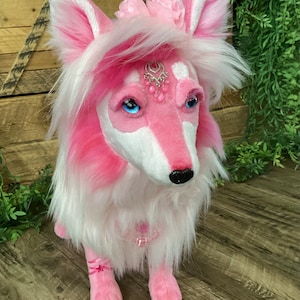 May include: Pink plush wolf with white fur, wearing a pink flower crown and a jeweled necklace. The wolf has blue eyes and a black nose.