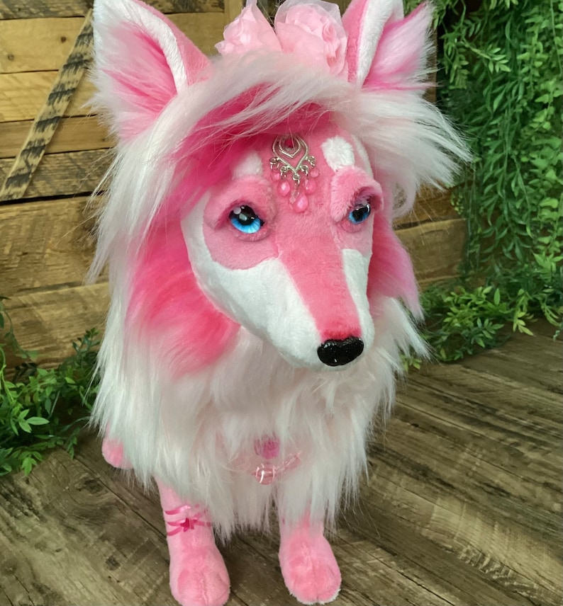 May include: Pink plush wolf with white fur, wearing a pink flower crown and a jeweled necklace. The wolf has blue eyes and a black nose.