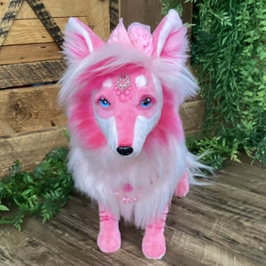 May include: A pink and white plush fox with blue eyes and a crown of pink flowers. The fox is wearing a pink and white necklace with a silver charm.
