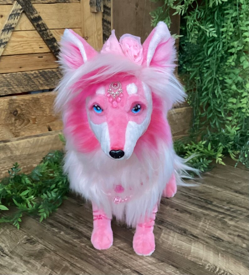 May include: A pink and white plush fox with blue eyes and a crown of pink flowers. The fox is wearing a pink and white necklace with a silver charm.