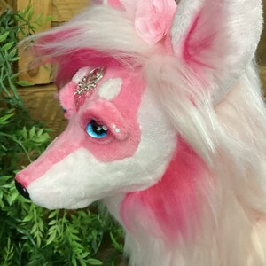 May include: A pink and white plush toy with long white hair, a pink flower on its head, and blue eyes. The toy has a silver and pink jeweled ornament on its forehead.