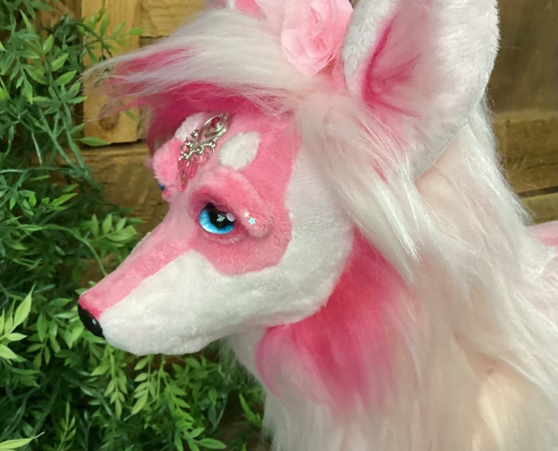 May include: A pink and white plush toy with long white hair, a pink flower on its head, and blue eyes. The toy has a silver and pink jeweled ornament on its forehead.