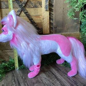 May include: A pink and white plush wolf with long white fur and a pink tail. The wolf is standing on a wooden floor.