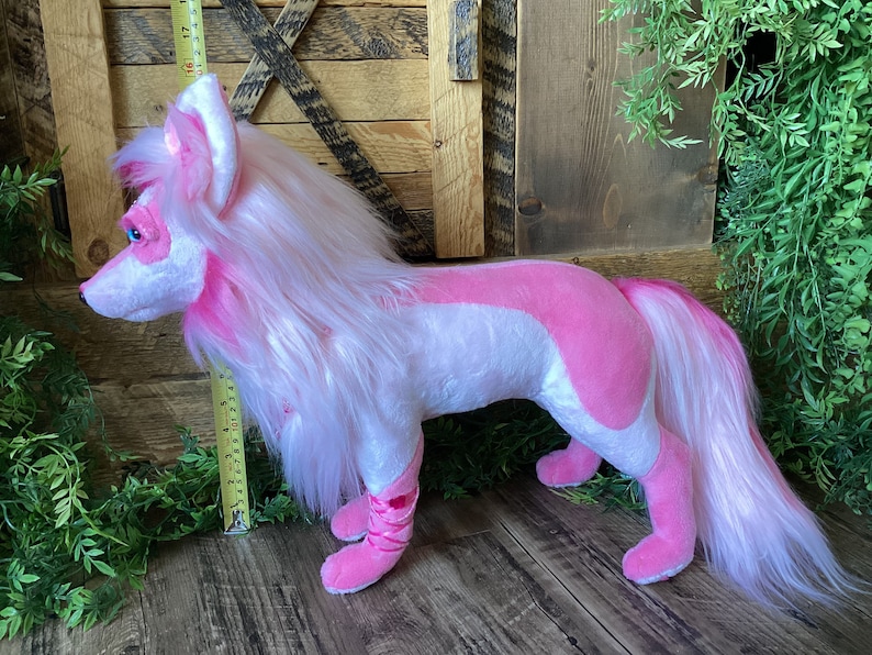 May include: A pink and white plush wolf with long white fur and a pink tail. The wolf is standing on a wooden floor.