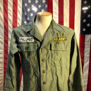 May include: A green military jacket with a name tag that reads "PALUMBO" and a patch that reads "U.S. ARMY".