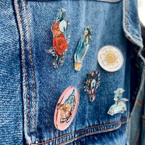 以下が含まれることがあります： デニムジャケットに、さまざまな色のピンが付けられています。ピンには、花柄、虎、太陽、ヤシの木、漫画のキャラクターが描かれています。