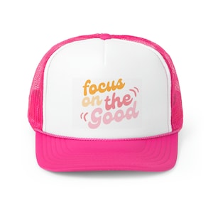 Peut inclure: Une casquette de camionneur blanche avec un filet rose à l'arrière. Le devant de la casquette présente un motif rose, orange et jaune qui dit "focus on the "good""