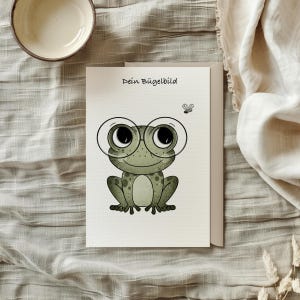 Könnte beinhalten: Eine weiße Karte mit dem Text "Dein Bügelbild" und einer Cartoon-Illustration eines grünen Frosches, der eine runde Brille trägt. Eine kleine Fliege ist über dem Frosch gezeichnet.
