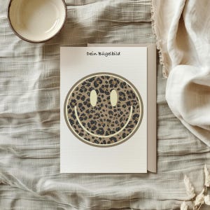 Könnte beinhalten: Eine Grußkarte mit einem Smiley-Gesicht im Leopardenmuster. Das Smiley-Gesicht hat einen beigen Rand und hellgelbe Augen. Der Text "Dein Bügelbild" ist oben auf der Karte aufgedruckt. Die Karte liegt auf einer neutralfarbenen Oberfläche.