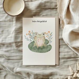 Könnte beinhalten: Eine weiße Karte mit dem Text "Dein Bügelbild" in schwarzer Schrift. Ein grüner Frosch mit rosa Bauch sitzt auf einem grünen Seerosenblatt mit orangen Blüten und grünen Blättern.