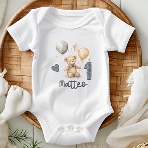 Könnte beinhalten: Weißer Baby-Strampler mit einem braunen Teddybär, der einen grauen Ballon mit der Nummer eins hält. Der Strampler hat den Namen Matteo darauf gedruckt.