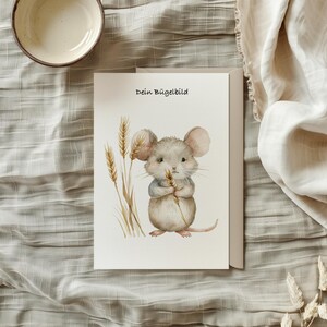 Könnte beinhalten: Eine weiße Karte mit dem Text "Dein Bügelbild" in schwarzer Schrift. In der Mitte der Karte befindet sich eine Aquarell-Illustration einer grauen Maus, die einen Weizenhalm hält. Die Maus steht auf einem grünen Stiel mit Weizenähren.