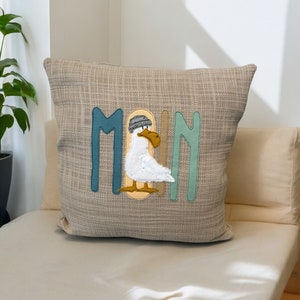 Könnte beinhalten: Ein beiger Kissenbezug mit dem Wort "Moin" in bunten Buchstaben bestickt. Ein weißer Seemann mit einer grauen Mütze befindet sich in der Mitte des Wortes.
