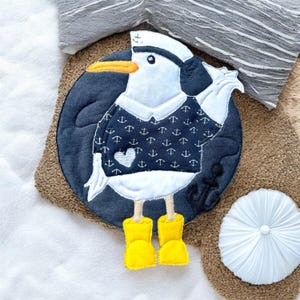 Könnte beinhalten: Eine runde Stoffapplikation in Blau und Weiß, die eine Möwe mit Matrosenhut und einem Pullover mit Ankern zeigt. Die Möwe trägt gelbe Gummistiefel.