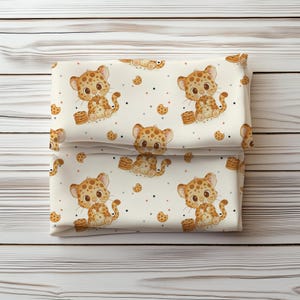 Könnte beinhalten: Gefalteter Stoff mit einem sich wiederholenden Muster aus Cartoon-Leopardenbabys und Keksen. Die Babys sind hellbraun mit braunen Flecken, und die Kekse sind braun. Der Hintergrund ist cremefarben mit kleinen Punkten.