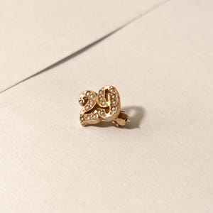 May include: A gold-colored pin with the number "29" is displayed on a white surface. The pin is adorned with small, round, pearl-like embellishments. The pin is likely a piece of jewelry or an accessory.