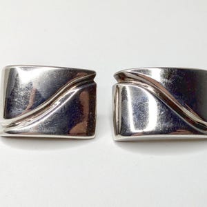 May include: Two silver-toned rectangular earrings. Each earring has a curved, wave-like design across the front. The earrings are reflective and appear to be made of metal. The earrings are displayed on a white surface.