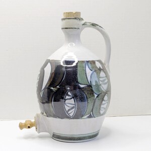 Puede incluir: Una jarra de agua de cerámica con tapón de corcho y grifo. La jarra presenta una base y un asa blancas, con un patrón decorativo de formas azules, verdes y negras. La jarra está diseñada para servir bebidas.