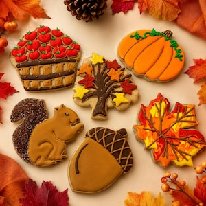 May include: A collection of six decorated sugar cookies featuring autumn themes: a basket filled with red apples, an orange pumpkin with green vines, a brown tree with colorful leaves, a brown squirrel, a brown acorn, and a large maple leaf.