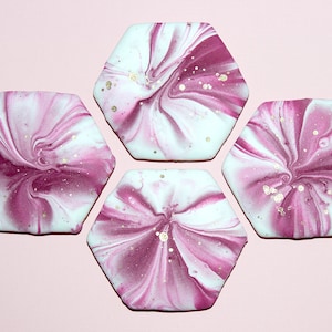 May include: Four hexagonal coasters featuring a marbled pattern of deep magenta and white swirls. Each coaster has a smooth, glossy finish with scattered flecks of metallic gold paint integrated into the swirling design across the surface.