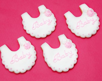 May include: Four white sugar cookies decorated with pink frosting in the shape of baby bibs. Each cookie has the word "Baby" written in pink frosting and a pink flower.