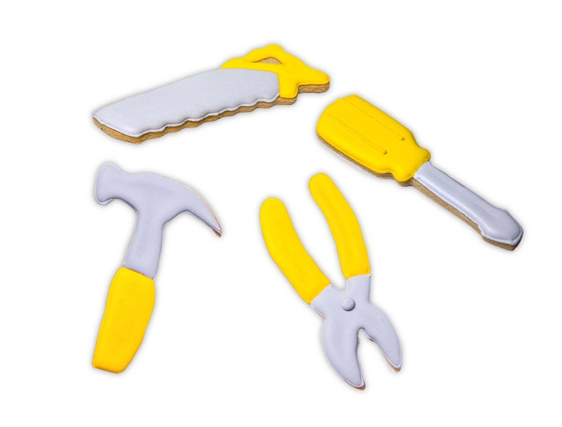 K&ouml;nnte beinhalten: Vier Werkzeug-f&ouml;rmige Kekse: eine S&auml;ge, ein Schraubendreher, ein Hammer und eine Zange. Die S&auml;ge und der Schraubendreher haben gelbe Griffe und graue Klingen. Der Hammer hat einen gelben Griff und einen grauen Kopf. Die Zange ist gelb und grau. Auf wei&szlig;em Hintergrund.