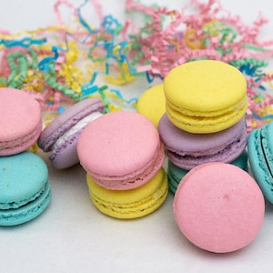 May include: A collection of pastel-colored macarons, including pink, yellow, blue, and purple, arranged on a white surface with pastel-colored shredded paper.