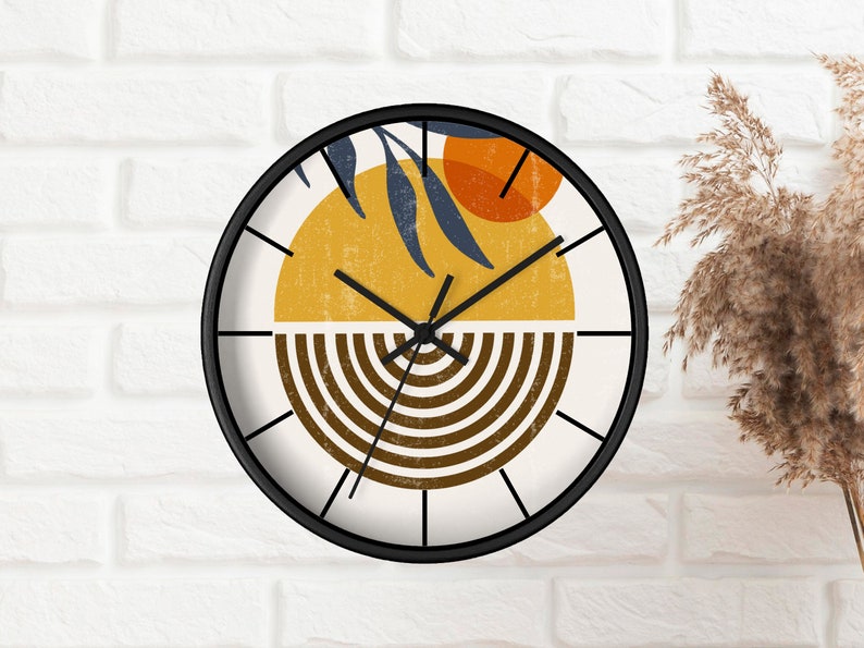 May include: A round clock with a black frame and white background. The clock face features a stylised design with a yellow sun, orange circle, and brown stripes. The clock hands are black.