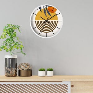 May include: A white clock with a brown, yellow and orange abstract design. The clock is hanging on a white wall above a wooden shelf with potted plants.