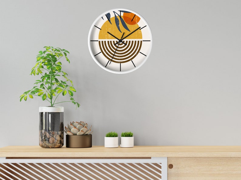 May include: A white clock with a brown, yellow and orange abstract design. The clock is hanging on a white wall above a wooden shelf with potted plants.