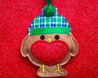 Könnte beinhalten: Ein Weihnachtsornament aus Filz in Form eines Rotkehlchens. Der Vogel hat einen braunen Körper, eine herzförmige rote Brust und eine grün-blau karierte Mütze mit einem grünen Bommel. Es hängt an einem grünen Band.