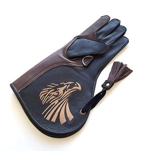Può includere: Un guanto in pelle marrone e nero con un design di aquila ricamato sul retro. Il guante ha un fiocco attaccato al polso.