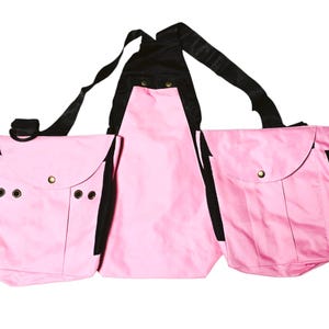 May include: Pink canvas bag with three compartments and black straps. The bag has a large central compartment and two smaller side compartments. The bag has a unique design with a strap that goes over the shoulders.