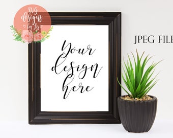 Könnte beinhalten: Ein schwarzer Bilderrahmen-Mockup mit dem Text "Your design here" auf weißem Hintergrund. Rechts im Bild befindet sich eine kleine grüne Topfpflanze.