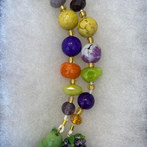 May include: A multi-strand necklace with green, yellow, orange, purple, and white glass beads. The necklace has a green pear and a purple grape charm at the end.