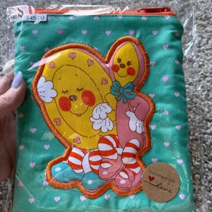 May include: A teal fabric pouch with a red zipper and a cartoon character applique. The applique features a yellow heart-shaped character and a pink character with a bow. The pouch is decorated with pink hearts and the words "homemade with love."