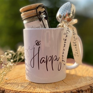 Könnte beinhalten: Weiße Keramiktasse mit dem Wort "Happy" in schwarzer Kursivschrift. Eine kleine Biene mit Flügeln fliegt über dem Wort. Die Tasse hat ein braunes Band um den Henkel gebunden, auf dem der Text "bling it on, match it" steht. Im Hintergrund befinden sich ein Glasgefäß mit Bambusdeckel und ein Metall-Tee-Infuser.