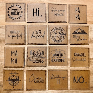 Op de afbeelding: Een verzameling vierkante, bruine leren patches met verschillende zwarte teksten en grafische ontwerpen. Zinnen zijn onder meer "Camp More Worry Less", "Hi", "Perfectly Imperfect" en "Over Dressed". Andere patches tonen berg- en kompasafbeeldingen.