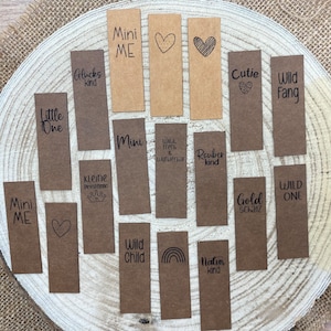 Könnte beinhalten: Eine Sammlung von 14 braunen Papier-Geschenk-Etiketten mit verschiedenen schwarzen Tinten-Designs und Texten. Der Text beinhaltet "Mini Me", "Wild Child", "Little One", "Wild Fang", "Cutie", "Gold Schatz", "Wild, frech & wunderbar", "Räuber kind", "Nature kind", "Kleine Prinzessin", "Glücks kind", and "Wild One".