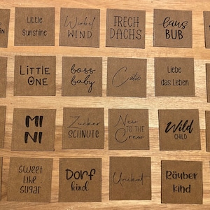 Può includere: Ventiquattro etichette di carta marrone con scritte in inchiostro nero. Le etichette presentano varie frasi e parole, tra cui "Little Sunshine", "Boss Baby", "Wild Child", "Sweet Like Sugar" e "Dorf Kind".