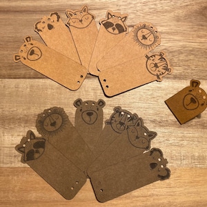 Può includere: Un set di 10 etichette regalo in carta marrone con varie illustrazioni di animali. Le etichette sono rettangolari e hanno dei fori perforati nella parte superiore per un filo o un nastro.