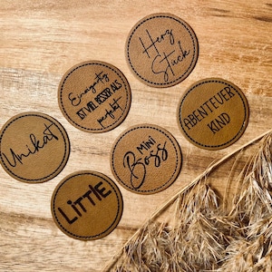 Può includere: Sei etichette rotonde in pelle marrone con testo nero. Le etichette dicono "Unikat", "Einzigartig ist viel besser als perfekt", "Herzstück", "Abenteuerkind", "Mini Boss" e "Little".