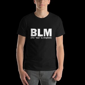 May include: Black t-shirt with the white letters "BLM" printed on the front. Below the letters, in smaller white text, it reads "(the "too" is implied)". The shirt is short-sleeved and appears to be made of a soft material.
