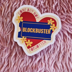 May include: A red heart-shaped sticker with the text "Gone But Not Forgotten" and a blue banner with the text "Blockbuster" in white. The sticker is decorated with yellow flowers with pink centres.