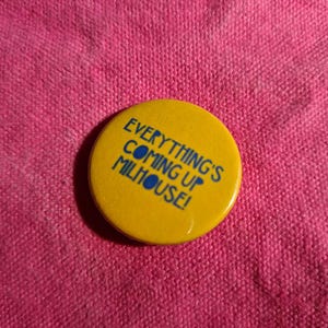 May include: A yellow button with the text "EVERYTHING'S COMING UP MILHOUSE!" in blue lettering. The button is circular and set against a pink fabric background. The button is a pinback button.