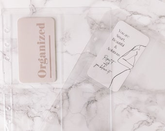 May include: Two clear plastic pockets for a planner or journal. The pockets are designed to hold cards or other small items. One pocket has a card with the word "Organized" printed on it. The other pocket has a card with the text "You are Smart, Beautiful & Ambitious. Repeat it until you believe it."